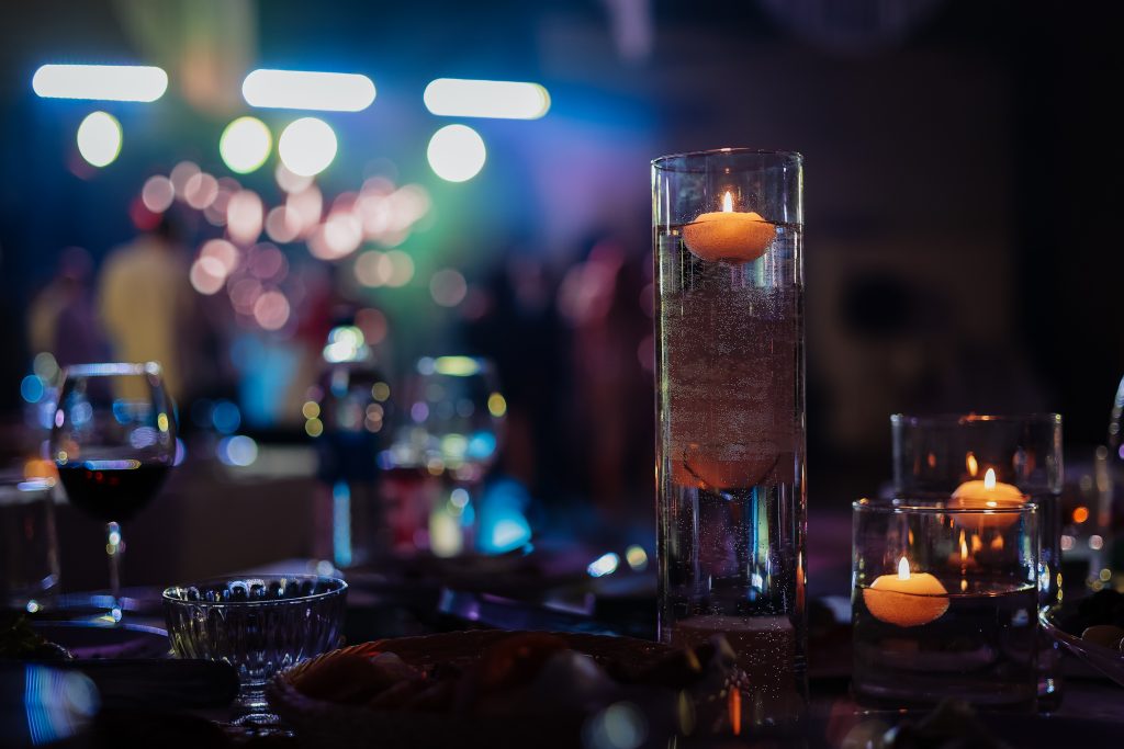 Banquet table decorated with burning candles in glass vases in restaurant hall. In the background party with silhouettes of people dancing on the dance floor with disco lights glowing searchlight.
